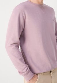 Mikina s dlouhým rukávem a kulatým výstřihem z jemné lila látky, s nenápadnou kapsou na prsou a žebrovanými manžetami. Textura působí hladce a pohodlně.