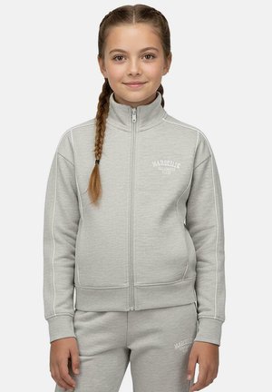 Meisje met gevlochten haar, gekleed in een lichtgrijs trainingspak met rits en de tekst "Marseille" op jas en broek, staand tegen een witte achtergrond.