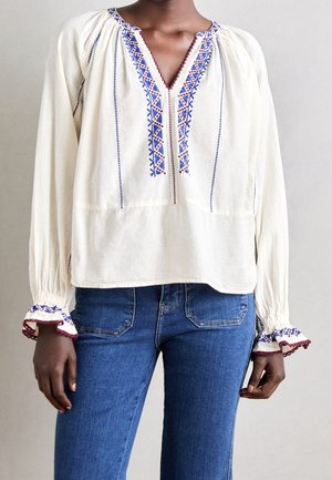 Personne portant un chemisier blanc avec des motifs géométriques brodés en bleu et bordeaux, associé à un jean denim bleu sur un fond uni.