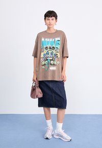 Brun oversized grafisk t-shirt med et farverigt Nike-design, parret med en mørk denimskørt og pastelfarvede sneakers, holdt med en lille pink punge.