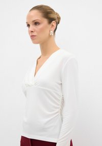 Top bianco a maniche lunghe con scollatura a V e cuciture laterali arricciate. Tessuto liscio e leggero, design aderente. Il modello indossa orecchini di argento oversize.