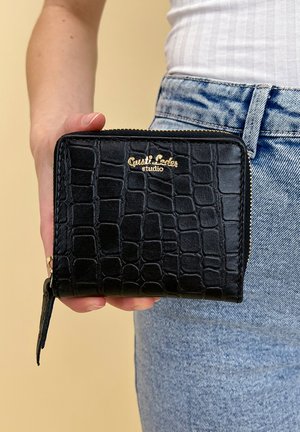 Schwarze Lederbörse mit strukturiertem Krokodilmuster, goldfarbenem Logo, Reißverschluss und kleinem Riemen. Wird in einer Hand vor einer Jeans gehalten.