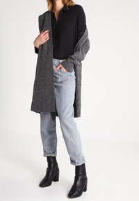 Camicia nera a bottone, cardigan grigio a maglia, jeans in denim chiaro e stivali neri con tacco alla caviglia. Tessuto testurizzato con vestibilità comoda e polsini.