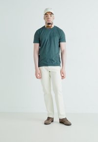 Mørkegrønn kortermet t-skjorte, kremfargede bukser og brune sko. Enkelt design, ensfargede, og avslappet passform. Lett caps på hodet.