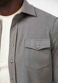 Harmaa nappikauluspaita, joka on valmistettu pehmeästä kankaasta. Paidassa on rintatasku, näkyvät saumat ja helmiäispintaiset napit edessä.