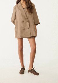 Blazer beige de doble botonadura con rayas verticales y mangas enrolladas. Combinado con una falda a juego y zapatillas de cordones marrones.