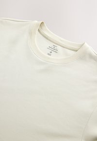 T-shirt écru taille moyenne, coupe régulière, col rond avec étiquette intérieure indiquant la taille et les détails du tissu.