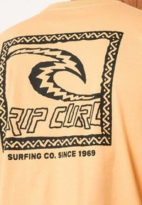 Lichtoranje t-shirt met gedurfde zwarte grafische ontwerpen met golven en de tekst "Rip Curl", omrand door een patroonrand. Katoenen materiaal.