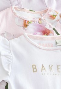 Tri majice za malu djecu: ružičasta s cvjetnim uzorkom, bijela s zlatnim natpisom "BAKER" i svijetloplava. Sve imaju okrugle izreze i nježne tkanine.