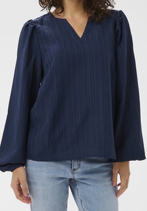 Person wearing a long-sleeved, navy blue V-neck blouse with subtle vertical stripes and light blue jeans, shown from shoulders to hips.