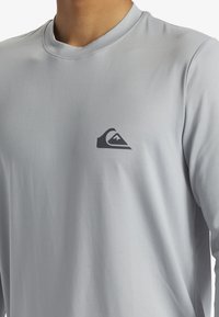 Svetlošedá košeľa s dlhým rukávom vyrobená z hladkého materiálu, s malým tmavým logom na ľavej hrudi a okrúhlym výstrihom.