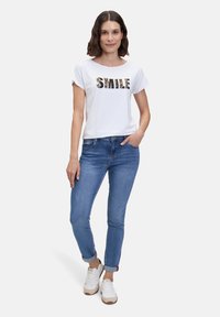 T-shirt blanche à manches courtes avec un imprimé « SMILE » en lettres colorées, associée à un jean slim bleu et des baskets blanches. Design simple.