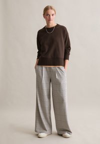 Brązowa bluza o luźnym kroju, zestawiona z szerokimi spodniami w szaro-białą kratę, wyróżniającymi się plisowanym fasonem i elastycznym paskiem w talii.