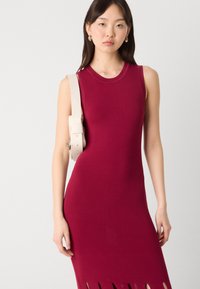 Czerwona sukienka bez rękawów o dopasowanym fasonie z wycięciami na dole. Posiada beżową torbę na pasku noszoną na krzyż na ramieniu.