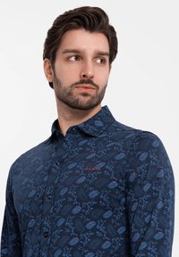 Marineblauwe knopshirt met een bloemenpatroon in lichtblauw en rood. Zachte textuur met een klassieke kraag en geborduurd logo op de borst.