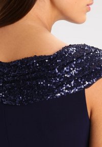 Marinblå klänning med en paljetterad off-shoulder halsringning. Paljetterna är glänsande och tätt packade, vilket kontrasterar med det släta tyget.