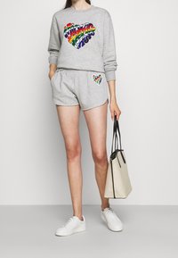 Felpa grigia con grafica a forma di cuore arcobaleno, abbinata a pantaloncini grigi coordinati con un piccolo dettaglio a forma di cuore. Il modello tiene una borsa beige. Sneakers bianche.