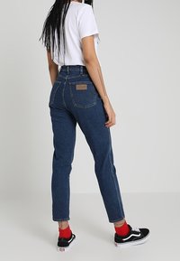 Žena s dlouhými copy, oblečená v bílém tričku, vysokých modrých džínách, červených ponožkách a černých teniskách, stojící na jednobarevném pozadí.