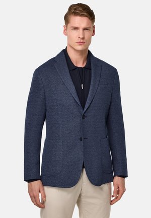 Blazer azul marino con tejido texturizado, dos bolsillos frontales, solapas de muesca y cierre de un solo botón, superpuesto sobre una camiseta polo oscura.