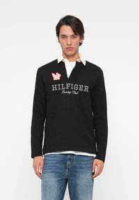 Melna ilga piedurkne polo krekls ar baltu apkakli, uz kura izšūts balts teksts "HILFIGER Racing Club" un sarkans izšūts logo uz krūtīm.