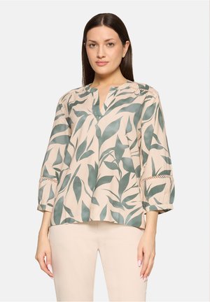 Frau mit glattem, dunklem Haar trägt beige Bluse mit grünem Blattmuster und cremefarbene Hose, steht vor einfarbigem Hintergrund.