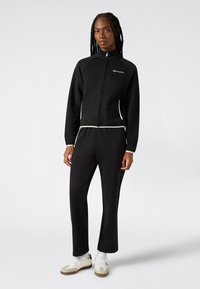 Czarny dres wykonany z miękkiego materiału, z zamkiem z przodu i kontrastującymi białymi akcentami na kieszeniach i mankietach, zestawiony z białymi trampkami.