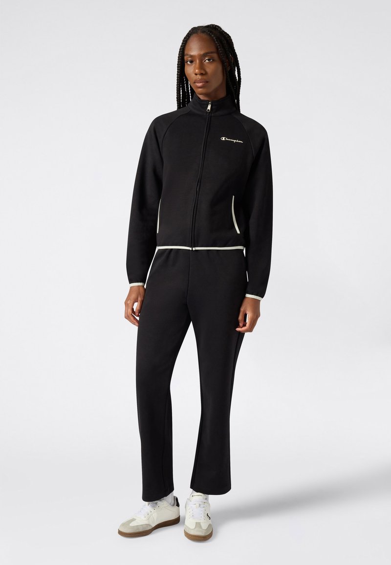 Czarny dres wykonany z miękkiego materiału, z zamkiem z przodu i kontrastującymi białymi akcentami na kieszeniach i mankietach, zestawiony z białymi trampkami.