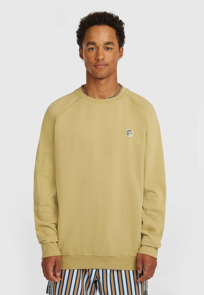 Bež jopica z okroglim ovratnikom, dolgimi raglanskimi rokavi in rebrastimi manšetami. Spredaj je okrogla značkasta nalepka na prsih.