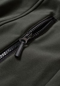 Zbliżenie na czarny zamek błyskawiczny na ciemnozielonym materiale, ukazujące zęby zamka i uchwyt z odblaskowym paskiem.