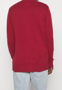 Czerwony sweter z dzianiny z prążkowaną fakturą, dekoltem w serek i długimi rękawami. Dół również jest prążkowany. Noszony z jasnoniebieskimi dżinsami.