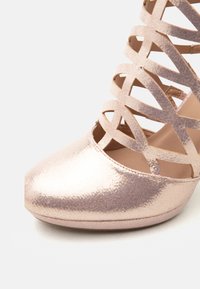 Λουστρινένιο παπούτσι με ψηλό τακούνι σε ροζ χρυσό χρώμα, με μυτερή μύτη και σχέδιο με σταυρωτά λουράκια πάνω από το πόδι και τον αστράγαλο.