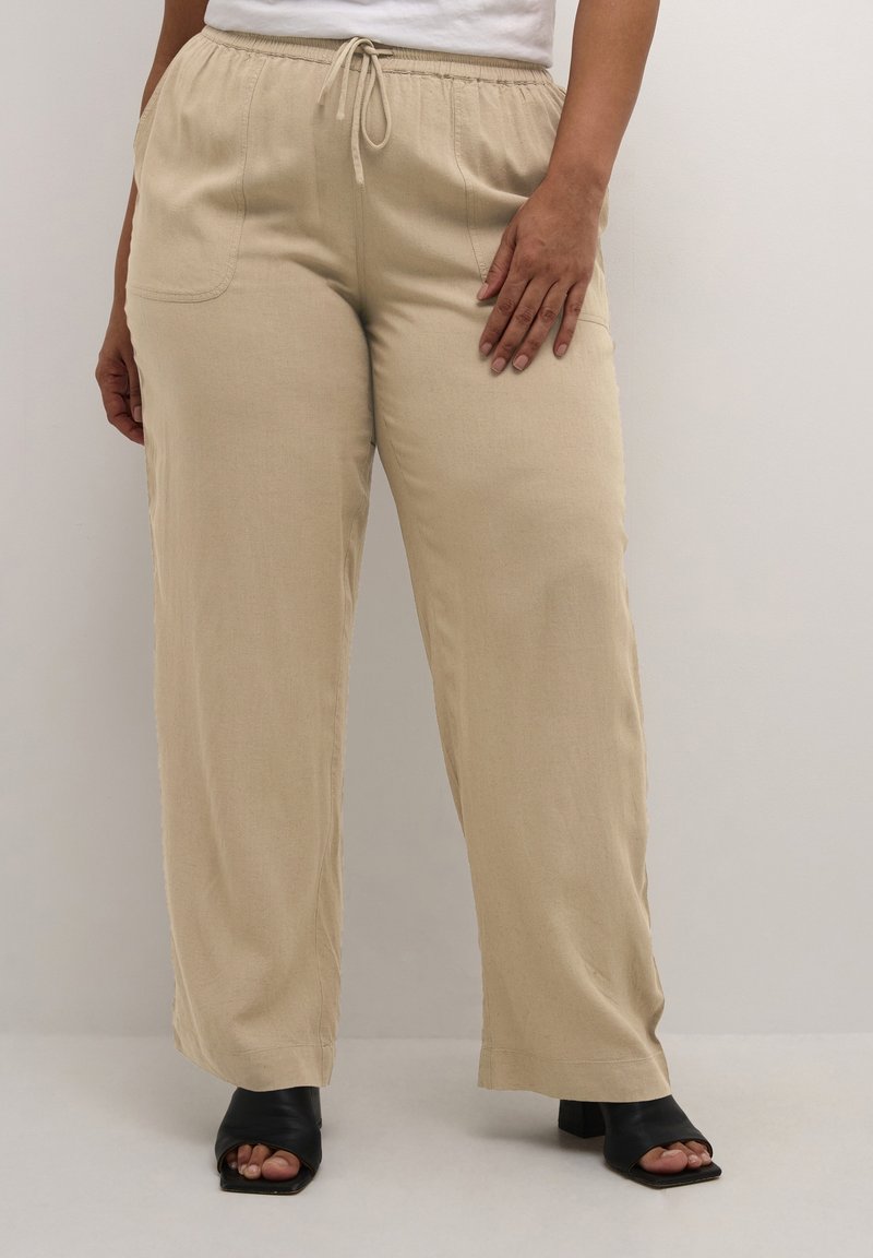 Bas du corps d'une personne portant un pantalon beige à cordon de serrage et des mules à talons ouvertes noires, avec une main reposant sur la cuisse contre un fond clair.