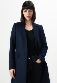 Frau mit glattem dunklem Haar, die einen marineblauen Mantel über einem schwarzen Oberteil und dunklen Jeans trägt. Sie steht mit einer Hand in der Tasche vor einem weißen Hintergrund.
