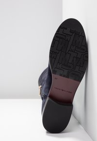 Suola di stivale alla caviglia in suede blu navy con cerniera, tacco in gomma nera texturizzata e logo "Tommy Hilfiger" impresso sulla suola marrone.