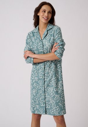 Femme souriante, bras croisés, portant une robe boutonnée bleu sarcelle avec des motifs floraux blancs et un col, sur un fond uni.