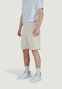 Pantaloni corti beige con vita elastica, dotati di un logo in rilievo discreto sul lato; abbinati a una maglietta bianca e a scarpe da ginnastica bianche.