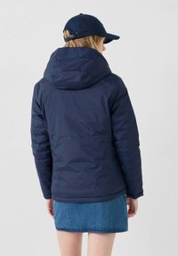 Giubbotto imbottito blu navy con cappuccio e texture trapuntata, abbinato a una gonna di jeans e un cappello coordinato. La vista posteriore mostra linee fluide.