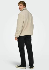 Beige fleece-takki korkealla kauluksella, teksturoidulla pinnalla ja pitkillä hihoilla, yhdistetty mustiin farkkuihin ja mustiin lenkkareihin.