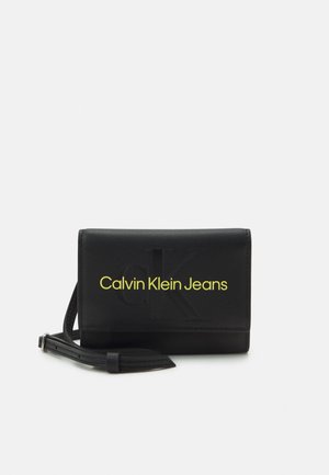 Portefeuille noir Calvin Klein Jeans avec logo "CK" en relief et nom de la marque en jaune, accompagné d'une sangle de poignet amovible.