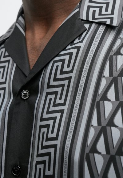 Κοντομάνικο πουκάμισο σε μαύρο και γκρι με γεωμετρικά σχέδια, που περιλαμβάνουν ζιγκ ζαγκ και κάθετες ρίγες, με μαύρο κούμπωμα με κουμπιά.