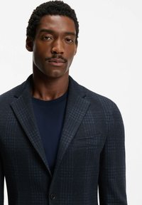 Mężczyzna z wąsami ubrany w ciemnoniebieską, kratkowaną marynarkę na granatowej koszulce z okrągłym dekoltem, zwrócony przodem, na białym tle.