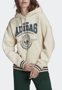 Persona che indossa un hoodie crema Adidas con logo, testo ed emblema, abbinato a pantaloni verde scuro, con le mani incrociate in vita.