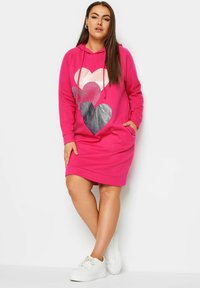 Vaaleanpunainen hupparitunika, jossa on sydänkuvio hopeanvärisenä ja mustana. Mallissa on taskut ja rento istuvuus, yhdistetty valkoisiin lenkkareihin.