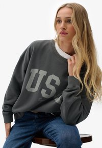 Grijze oversized sweatshirt met "US" in grote letters gedrukt. Heeft een ronde hals en geribbelde manchetten, gestyled over een wit shirt.