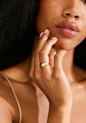 Anillo de oro con un diseño liso y redondeado que se posa en un dedo, con un acento en forma de corazón. La mano toca suavemente el rostro.