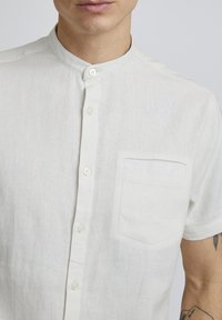 Chemise en lin blanche à manches courtes, avec col montant, fermeture à bouton sur le devant et une poche poitrine. Texture lisse et design léger.