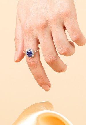 Bague en argent ornée d'une pierre bleue ovale, entourée de petites pierres claires, portée sur un doigt devant un fond doux et clair.