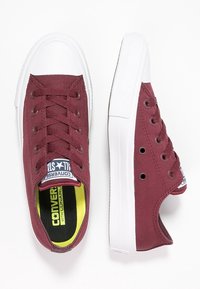 Para bordowych niskich tenisówek z płótna z białymi gumowymi podeszwami, białymi czubkami palców i białymi sznurówkami, widziana z góry i z boku.