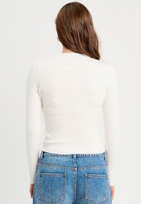 Pull en maille côtelée blanche à manches longues, texture détaillée, design ajusté ; associé à un jean taille haute bleu clair, vue de dos.
