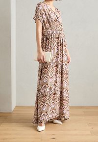 Robe maxi en tissu à motifs présentant des formes de diamant en marron, rose et vert. Matériau doux et fluide avec des manches courtes. Chaussures blanches.